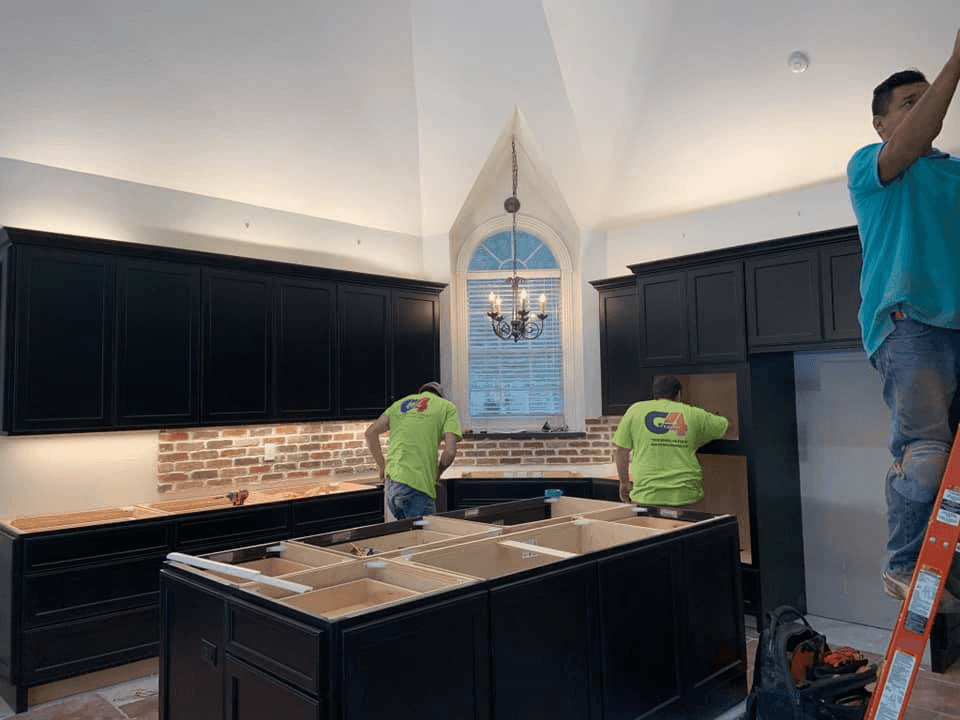 Workers installing cabinets in a modern kitchen with a brick backsplash and chandelier.